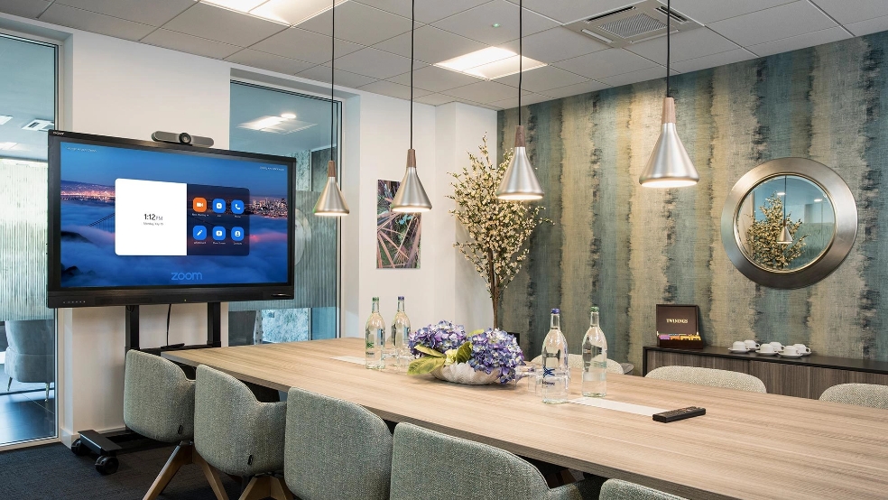 Meeting room with a long wood table and grey chairs, with a conference tv