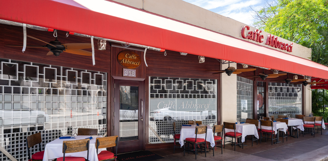 Photograph of the front of Caffe Abbracci, its entrance is made of wood and has several chairs and tables outdoors.