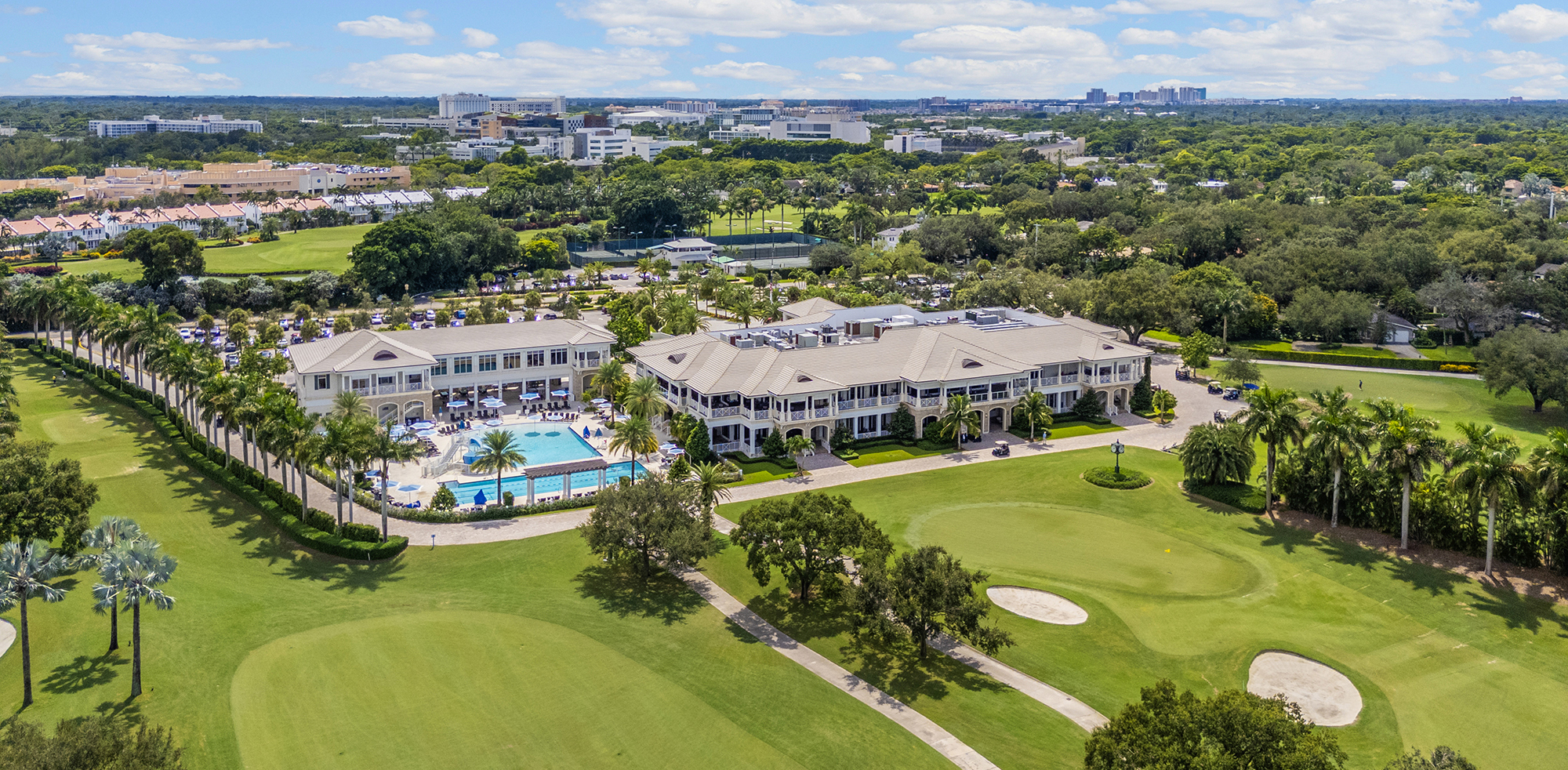 Photograph of a gray hotel with a pool in the middle, surrounded by trees, bushes, and some buildings.