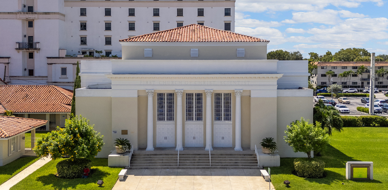 Front view of a beige building with four columns, a red-tiled roof, three white doors, and stairs leading to the entrance. Trees and green grass are in the foreground.