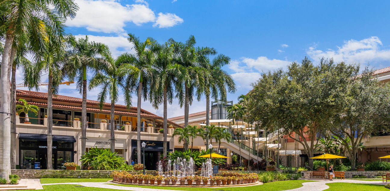 Outdoor shopping center with palm trees, boutique storefronts, and a water fountain at The Shops at Merrick Park in Coral Gables, Florida.