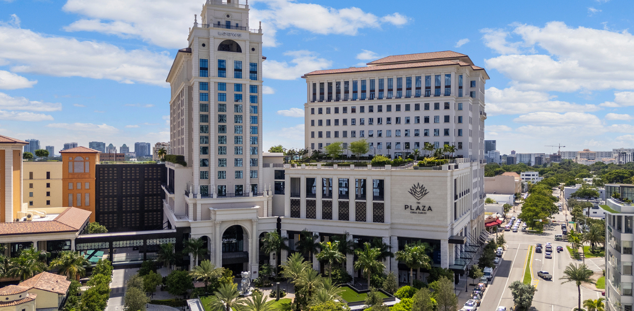 A large, multi-story building complex with a tower, labeled “PLAZA,” stands along a city street lined with palm trees under a blue sky with scattered clouds.